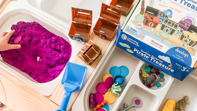 a toddler's hands digging in purple sand with a blue shovel with treasure chest toys nearby